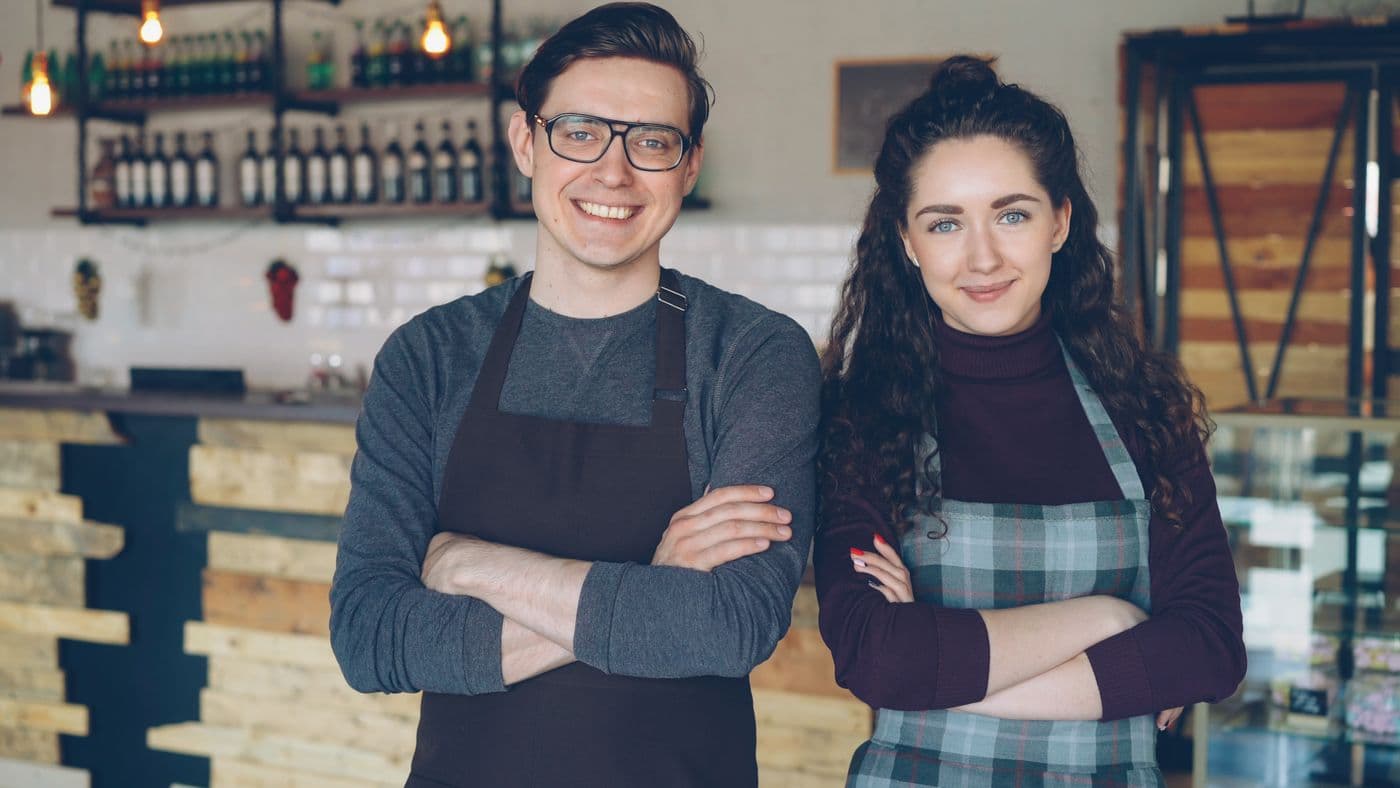 Two café owners in aprons standing in their coffee shop